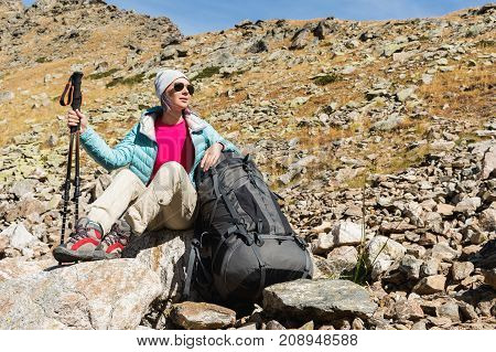 A tourist girl wearing sunglasses down jacket and hat with a backpack and mountain equipment with handles for tracking in her hands is sitting and relaxing looking at the beautiful view of the high mountain lake in the North Caucasus. Karachay-Cherkess