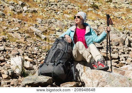 A tourist girl wearing sunglasses down jacket and hat with a backpack and mountain equipment with handles for tracking in her hands is sitting and relaxing looking at the beautiful view of the high mountain lake in the North Caucasus. Karachay-Cherkess 