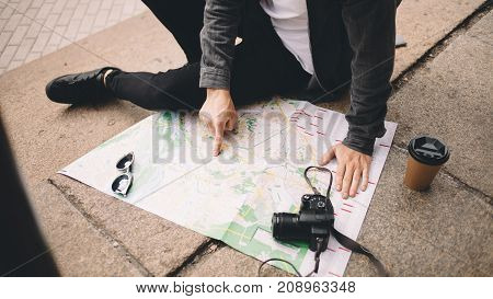 Close view of a young hipster that likes to travel. He decided to have some rest and sit down on the steps on the street. His coffee is getting colder while the guy is studing the map of the city. Close up. Cut view