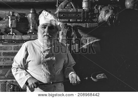 Senior male chief cook bearded man in uniform holds ash coal scoop and stands on kitchen background