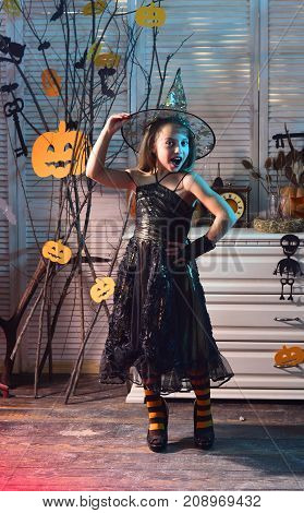 Halloween party and celebration concept. Little witch with Halloween decor. Girl with cheerful face on spooky carnival room background. Kid in witch hat and costume poses on high heels