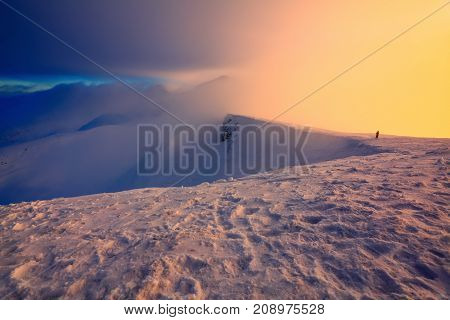 At the edge of the snowy rock someone is standing. The high mountains in the fog morning sky is enlighten with the orange colored light in winter day.