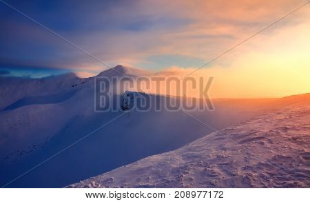 At the edge of the snowy rock someone is standing. The high mountains in the fog morning sky is enlighten with the orange colored light in winter day.