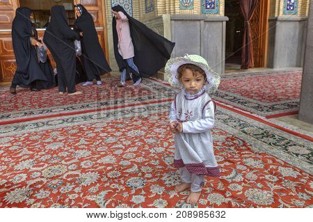 Shiraz Iran - 19 april 2017: Shah Cheragh Shrine Portrait of a unknown little girl about 4 years old standing in courtyard of the mosque on the background of women wearing muslim black chador.
