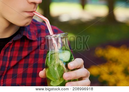 Young unrecognizable man drinks healthy detox tea with juice on green park background. Superfoods, health, summer freshness, vegetarian food concept