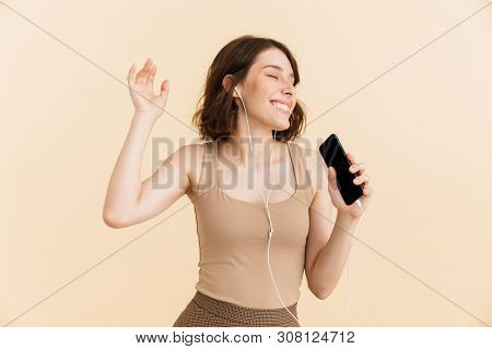 Portrait of optimistic young woman dressed in casual clothes singing while listening to music with earphones and cellphone isolated over beige background in studio