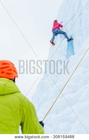Alpinist Woman With Ice Tools Axe In Orange Helmet Climbing A Large Wall Of Ice. Outdoor Sports Port