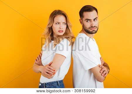Image of upset couple man and woman in basic t-shirts frowning while standing back to back during fight isolated over yellow background