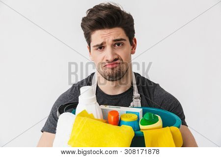 Handsome brunette houseman wearing apron standing isolated over white background, holding bucket full of detergents
