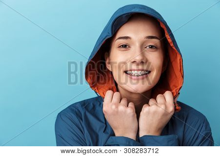Cheerful young girl wearing raincoat standing isolated over blue background, posing