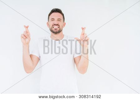 Young handsome man wearing casual white t-shirt over isolated background smiling crossing fingers with hope and eyes closed. Luck and superstitious concept.