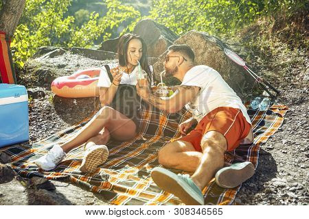 Young Couple Having Picnic At Riverside In Sunny Day. Woman And Man Spending Time On The Nature Toge