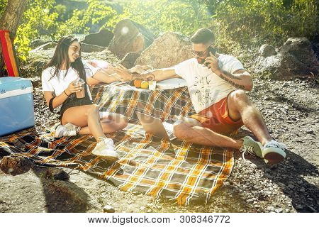 Young Couple Having Picnic At Riverside In Sunny Day. Woman And Man Spending Time On The Nature Toge