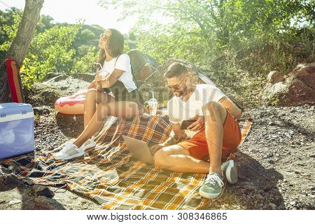 Young Couple Having Picnic At Riverside In Sunny Day. Woman And Man Spending Time On The Nature Toge