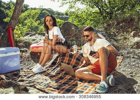 Young Couple Having Picnic At Riverside In Sunny Day. Woman And Man Spending Time On The Nature Toge