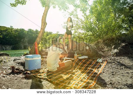 Young Couple Having Picnic At Riverside In Sunny Day. Woman And Man Spending Time On The Nature Toge