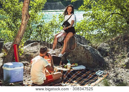 Young Couple Having Picnic At Riverside In Sunny Day. Woman And Man Spending Time On The Nature Toge