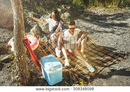 Young Couple Having Picnic At Riverside In Sunny Day. Woman And Man Spending Time On The Nature Toge