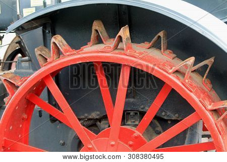 Horizontal Detail Of Antique Farm Equipment Tractor Wheel With Red Spokes And Rusty Metal Cleats For