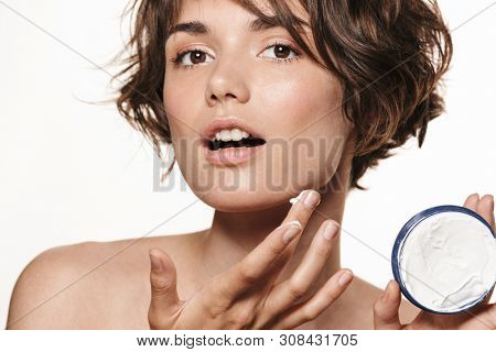Beauty portrait of a lovely young topless woman with short brunette hair standing isolated over white background, applying face cream, showing container, showing fingers