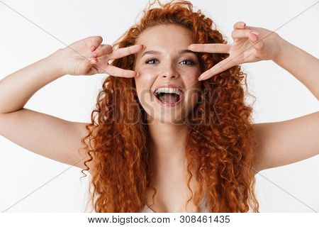 Close up portrait of an attractive joyful young woman with long curly red hair standing isolated over white background, peace geture