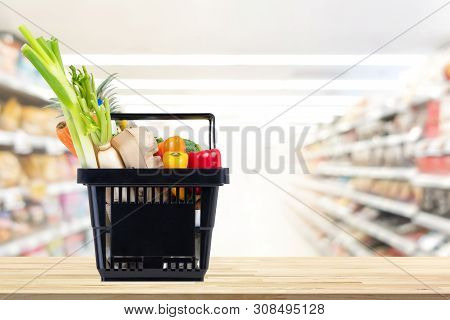 Food And Groceries In Black Shopping Basket On Wood Table With Blurred Suppermarket Aisle In Backgro