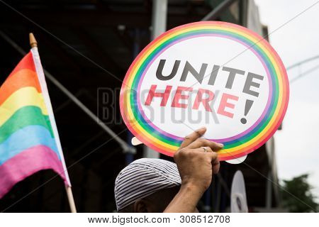 A hand holds a sign that says Unite Here with a colorful rainbow flag with building in background.