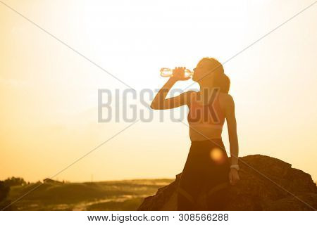 Young Woman Resting and Drinking Water after Trail Running Outdoor. Workout Exercise at Hot Beautiful Summer Sunset. Sport and Healthy Active Lifesyle Concept.