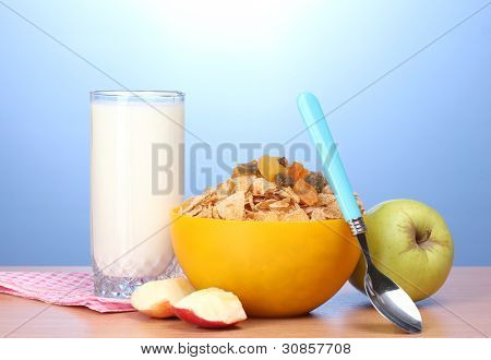 leckere Cornflakes in gelbe Schale, Äpfel und Glas Milch auf Holztisch auf blauem Hintergrund