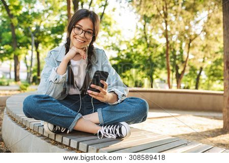 Photo of a pleased positive happy cute young student girl wearing eyeglasses sitting on bench outdoors in nature park using mobile phone chatting listening music with earphones.