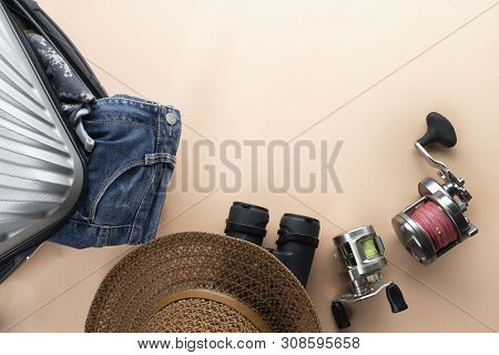 Flat Lay Grey Suitcase With Binoculars, Hat, Jeans, Spinning For Fishing And Sandles On Pastel Backg