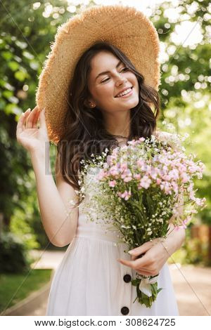 Image of excited happy young pretty woman outoors in green park walking in hat holding flowers.