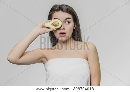 Young Astonished Girl With Half Avocado Is Looking Leftward. She Wears White Towel.