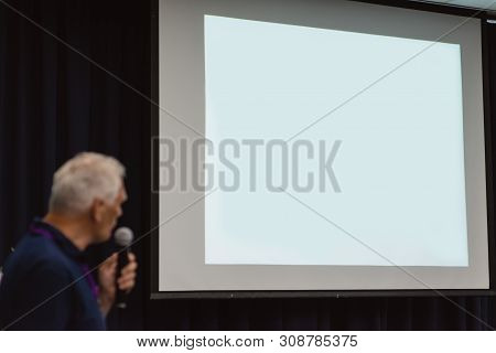 A Big White Screen Without Text. A Conference Concept. A Speaker With A Microphone Is Standing And W