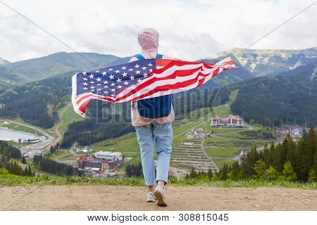 Back View Of Travel Standing At Top Of Hill Holding Usa Flag On Her Back, Wearing Jeans, Sneakers An