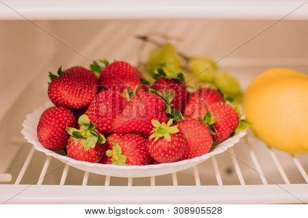 Plate With Ripe Strawberries Is On The Shelf Of The Refrigerator, Next To A Lemon And Grapes. The Co