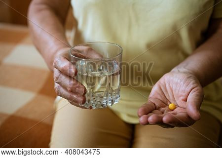 Mature Woman With A Glass Of Water Holds A Tablet In Her Hand While Sitting On The Couch. Treatment 