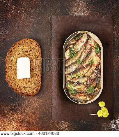 Open Tin Of Baltic Sprats In Oil With Spices, A Slace Of Bread With Butter On An Old Brown Backgroun