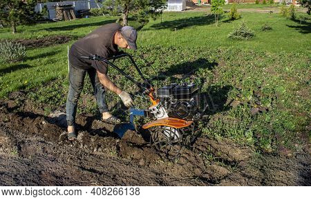 Male In The Spring Plows The Earth In Working Clothes With A Motoblock, Walk-behind Tractor