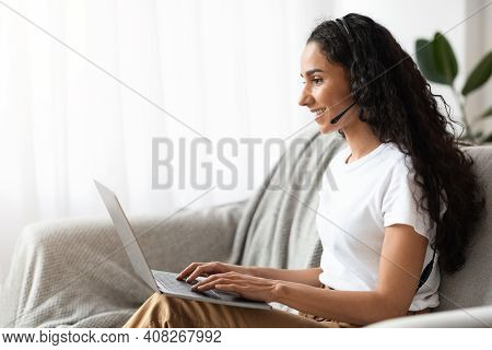 Side Vire Of Happy Pretty Woman With Dark Curly Hair Having Video Call From Home, Using Headset And 