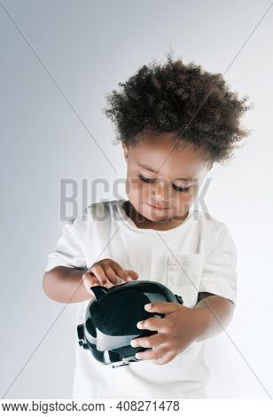 Portrait of a Nice African American Child Putting Coins in to the Pig-shaped Money Box Isolated on Grey White Background. Investment in to the Future