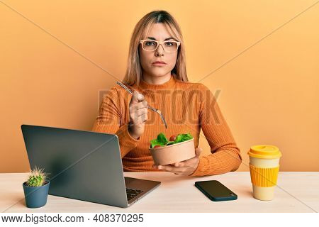 Young caucasian woman working at the office eating healthy salad relaxed with serious expression on face. simple and natural looking at the camera. 