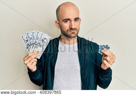 Young hispanic man playing poker holding dollars banknotes and chips relaxed with serious expression on face. simple and natural looking at the camera. 