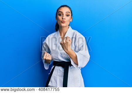 Beautiful brunette young woman wearing karate fighter uniform with black belt doing attack pose looking at the camera blowing a kiss being lovely and sexy. love expression. 