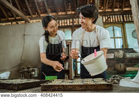 Smiling Asian Couple Wearing An Apron Looking Each Other While Pouring Dough On A Cake Pan
