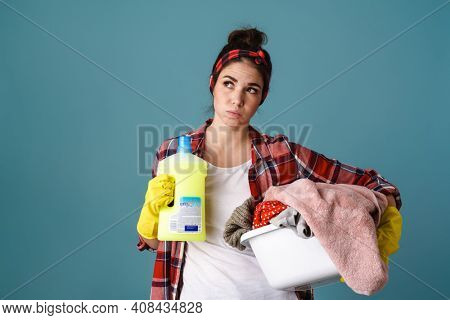 Upset housewife in apron and rubber gloves holding cleaning tools isolated over cblue