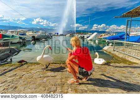 Blonde Tourist Girl Feeding White Swans On The Shore Of Geneva Lake In Geneva Harbor With 140m High 