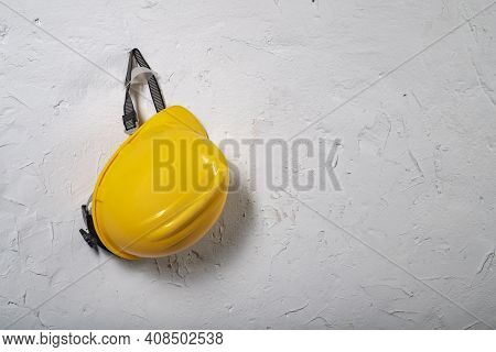 Yellow Work Helmet Hung On A Hook. Health And Safety Accessories On A White Wall Background.