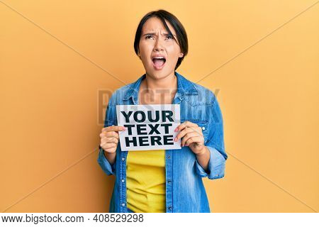 Beautiful young woman with short hair holding your text here message angry and mad screaming frustrated and furious, shouting with anger looking up. 