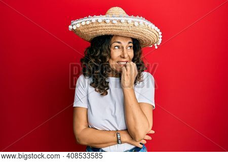 Middle age hispanic woman holding mexican hat looking stressed and nervous with hands on mouth biting nails. anxiety problem. 